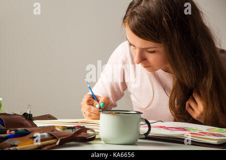 A young dark-haired artist paints in an album for drawing a thin wooden brush with watercolor, on the table brushes and a mug with tools Stock Photo