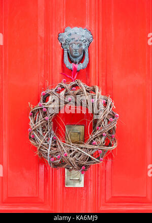 Natural Christmas wreath hanging from an ornate knocker on a bright red door. Stock Photo