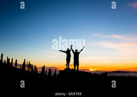 Teamwork couple climbing and reaching mountain peak. Silhouette of climbers team over mountains sunset. Man and woman hikers looking at inspirational  Stock Photo