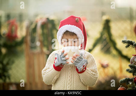 Beautiful school child, boy, holding Christmas mug, drinking tea in the garden, next to a Christmas tree on a frosty morning, smiling at camera Stock Photo