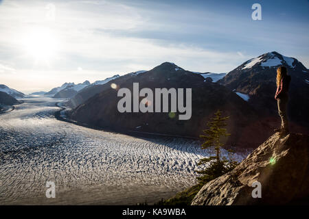 A young, curly haired blonde woman looks over the Salmon Glacier  in late evening golden light. A sun flare crosses the frame and many crevasses can b Stock Photo