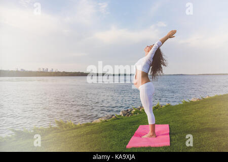 Young pregnant woman making yoga exercise and stretching outdoor. A woman standing on a yoga mat, raising her hands. River in the background Stock Photo