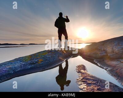 Tourist guy taking pictures of amazing sea landscape on mobile phone digital camera. Hiker stay above mirroring pool  on a rock near the sea level. A  Stock Photo