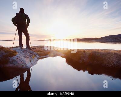 Tall backpacker watch clear sunny spring daybreak over sea. Hiker with backpack stand on rocky shore and his figure is mirrored in water pool.  Hiker  Stock Photo