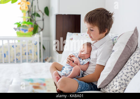 Sweet preschool boy, reading a book to his newborn brother, sitting on the bed in bedroom, childhood concept Stock Photo