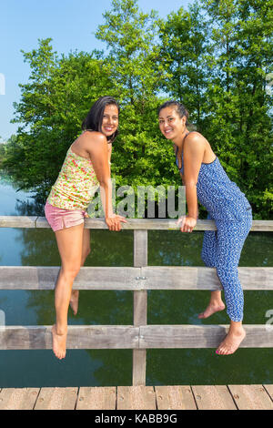 Two young women sitting together on bridge railing at water Stock Photo