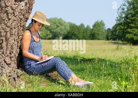 Young woman sitting against tree trunk writing in meadow Stock Photo