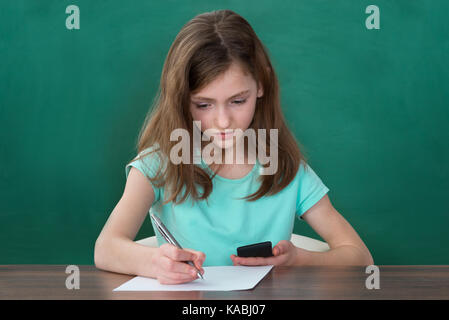 Girl With Mobile Phone Writing On Paper At Table In Classroom Stock Photo