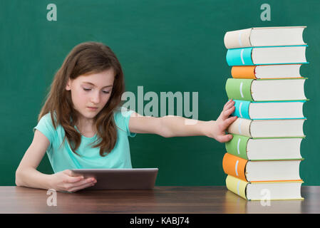 Girl Using Digital Tablet And Avoiding Stack Of Books At Desk In Classroom Stock Photo
