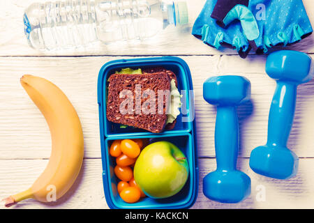 Toned photo of apple, sandwich, tomato in box, dumbbell, banana, water bottles on brown wooden table Stock Photo