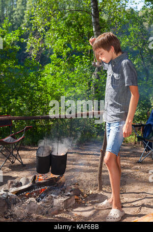 Young boy cooking camp food in cauldron on open fire Stock Photo