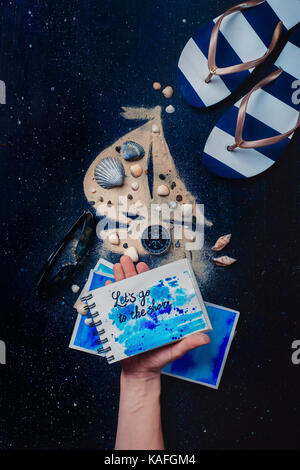 Sailboat silhouette made from sand and seashells, a notebook in a artist hand with handwritten text Let us go to the shore. Cocneptual still life with Stock Photo