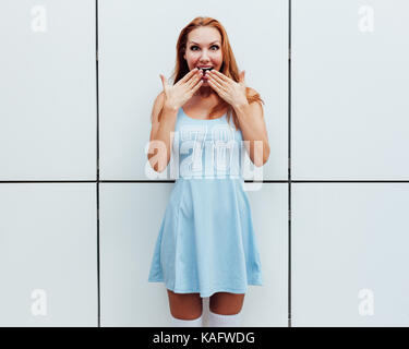 Surprise. Shock. Shocked and surprised girl screaming covering mouth her hands .Redhead young woman amazed. Beautiful girl in blue summer dress. Presenting your product. Expressive facial expressions. Outdoor. Stock Photo