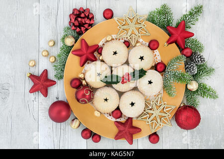 Homemade christmas mince pies on a gold plate with red and gold bauble decorations, holly, fir and foil wrapped chocolates on distressed white wood ba Stock Photo