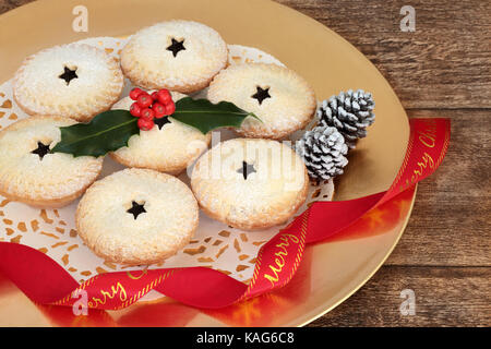 Christmas mince pies with holly and red ribbon on a gold plate on oak wood background. Stock Photo