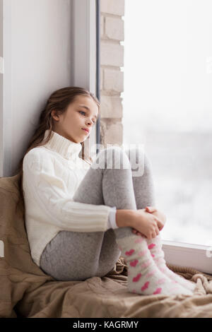 sad girl sitting on sill at home window in winter Stock Photo