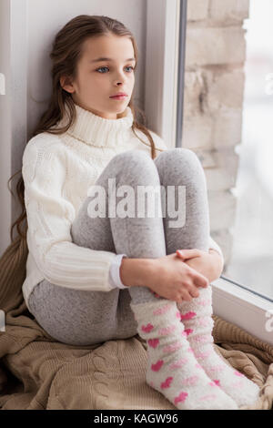 sad girl sitting on sill at home window in winter Stock Photo