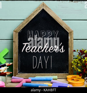a house-shaped chalkboard with the text happy teachers day written in it on a rustic wooden table full of pieces of chalk of different colors Stock Photo