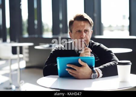 Portrait of thoughtful worried man sitting in cafe with digital tablet and biting glasses Stock Photo