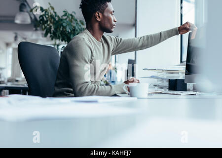 Young african man putting post-it note on computer monitor. Black male working at his desk in office. Stock Photo