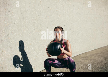 Sportswoman doing squat exercises with fitness ball. Female exercising and stretching with medicine ball outdoors. Stock Photo