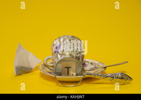 teabag with cup and saucer and silver spoon with letter T tile magnified with magnifying glass lens on yellow background. Love tea still life concept Stock Photo