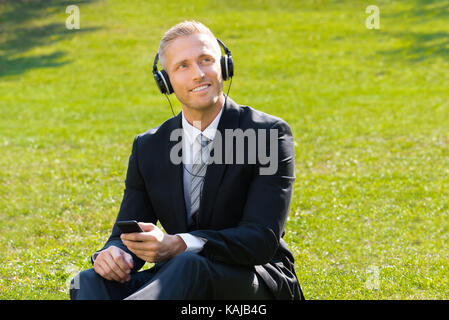 Young Smiling Businessman Sitting On Grass Listening Music In Park Stock Photo