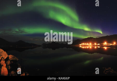 Green northern lights in night starry sky. Northern lights reflect in lake in small norwegian resort town. Beautiful north night lanscape. Stock Photo