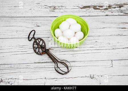 Antique eggbeater and a bowl of white eggs with a weathered wooden boards backdrop cut out antique images, US tools, vintage kitchen utensils Stock Photo