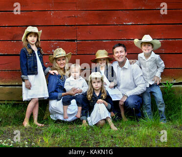 Family of seven pose besides red, wooden barn.  Females are wearing denim jackets and cowboy hats.  Males are wearing jeans and white shirts. Stock Photo