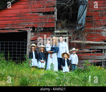 Father and mother pose with their five children in front of a rustic, red wooden barn.  Girls are wearing denim jackets and boys are wearing white shi Stock Photo
