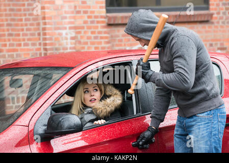 Male robber holding baseball bat while looking at scared woman in car Stock Photo