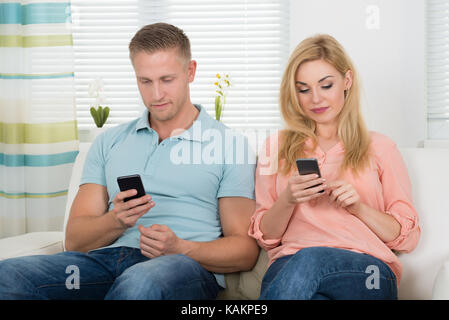 Young couple text messaging through mobile phones on sofa at home Stock Photo