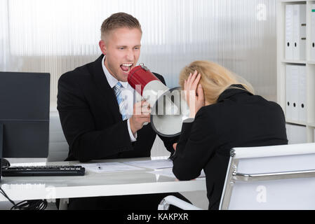 Boss shouting at businesswoman through loudspeaker at desk in office Stock Photo