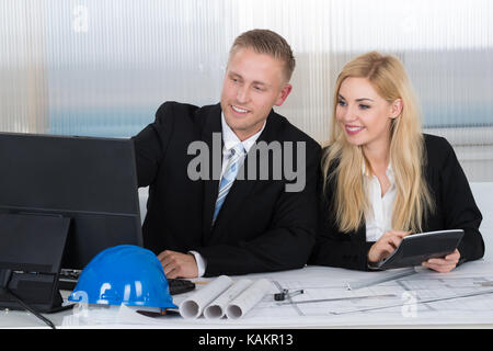 Young architects discussing over computer with blueprints on desk in office Stock Photo