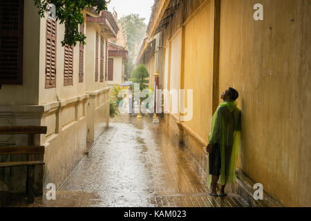A teenager wearing a transparent green rain jacket, waiting for the heavy monsoon rain to stop during a heavy rain shower in Phnom Penh city, Cambodia. Stock Photo