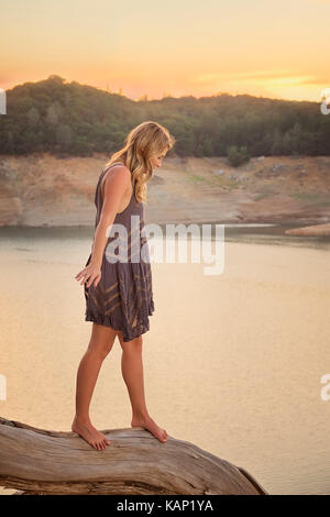 A young woman in a bohemian style dress balancing on a log above a mountain lake at sunset. Stock Photo