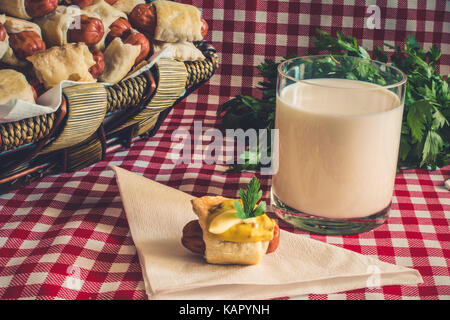 Wicker basket with artisan mini hot dogs (Sausage in the dough) with sausage on a napkin covered in mustard, mayonnaise and a glass of fresh cow's mil Stock Photo