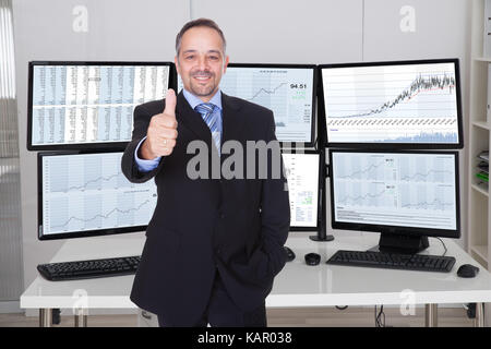 Portrait of stock market broker gesturing thumbs up against multiple monitors in office Stock Photo