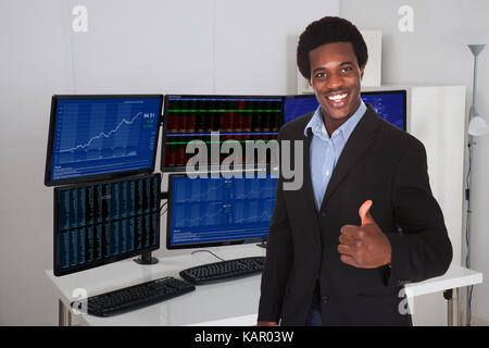Portrait of stock broker gesturing thumbs up against multiple monitors in office Stock Photo