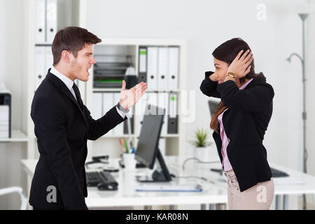 Side view of boss yelling at secretary covering ears in office Stock Photo