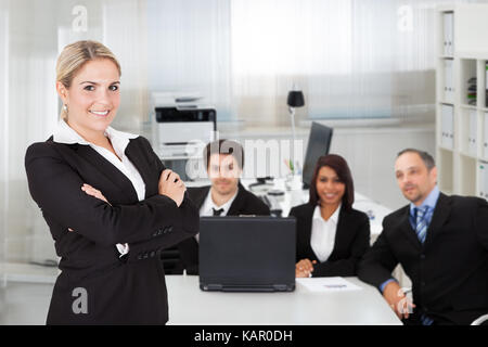 Portrait of young businesswoman standing arms crossed while colleagues sitting at desk in office Stock Photo