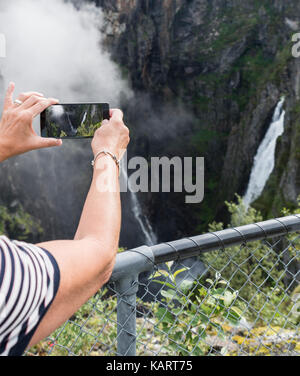 woman making photo with mobile phone of the Voringfossen waterfall in norway seen from street level on sunny day Stock Photo