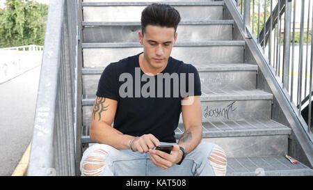 Handsome young man standing outdoors in urban environment on metal stairs, using cell phone typing message Stock Photo