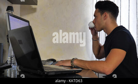 Attractive Young Man with Serious Expression, with Laptop and Desktop Comptuers, Very Busy Working on his Start-up Business Drinking Espresso Coffee - Stock Photo