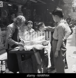 1950s, historical picture from Hong Kong of a female chinese food vendor, young boys stand by a street food stall looking at an elderly lady cooking on her small stove. Stock Photo