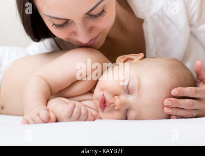 Beautiful young mom looking at her sleeping little baby and smiling. Child lying in bed at home Stock Photo