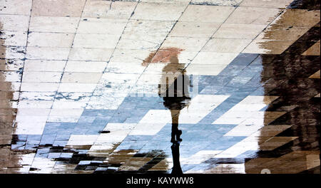 Blurry reflection shadow silhouette of a single woman under red umbrella walking in the old city pedestrian zone just after the rain Stock Photo