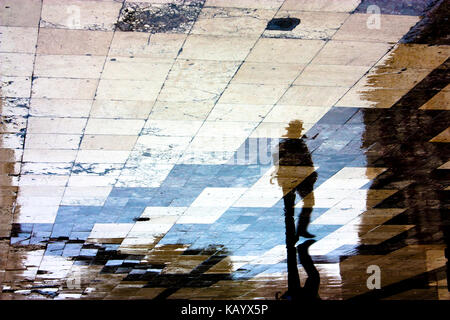 Blurry reflection shadow silhouette of a single man walking in the old city pedestrian zone just after the rain, with hat and folded umbrella Stock Photo