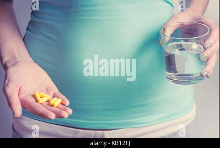 A pregnant woman first trimester in a blue T-shirt holds several medicine capsules, and in the second a glass of water. IVF Stock Photo